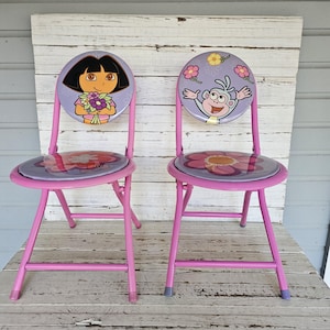 May include: Two pink folding chairs with round seats and backs. The seat and back of each chair feature a cartoon character and floral design. The chairs are made of metal and have a simple, functional design.