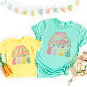 May include: Two yellow and turquoise t-shirts with a retro-style graphic of colorful Peeps and the text "Hello Peeps".
