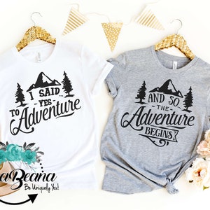 May include: Two t-shirts with a mountain and trees design. The white t-shirt says "I said... yes... to Adventure". The gray t-shirt says "And so... the Adventure begins".