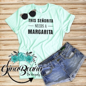 Op de afbeelding: Een lichtgroen T-shirt met de tekst "This Señorita Needs a Margarita" in zwart gedrukt. Het T-shirt wordt gecombineerd met een spijkerbroekje en een zonnebril.