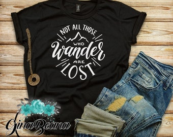 以下が含まれることがあります： 白いグラフィックが「Not all those who wander are lost」と書いてある黒いTシャツ。山がシルエットになっていて、白い縁取りがされている。