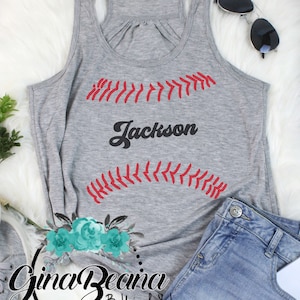 May include: Gray tank top with a red baseball stitch design and the name "Jackson" in black lettering. The tank top is on a white fuzzy surface.