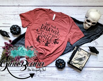 May include: A red t-shirt with the text "Just took a DNA test: Turns out I'm 100% witch" and a graphic of three witches flying on broomsticks. The shirt is on a white wooden background with black pumpkins and a black crow.