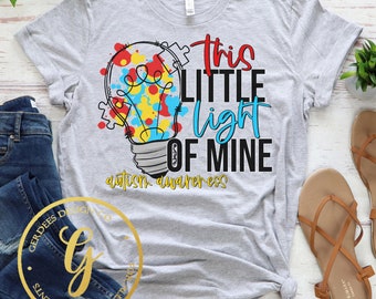 May include: Gray t-shirt with a light blue light bulb graphic with puzzle pieces and colorful paint splatters. The text "This little light of mine" is written in black with the words "autism awareness" below.