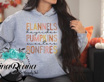 May include: Grey hooded sweatshirt with the text "FLANNELS hayrides PUMPKINS sweaters BONFIRES" in orange, yellow, and red. The text is layered and has a watercolour effect. A teal floral design is below the text. The text "Gina Beana Be Uniquely You!" is below the floral design.
