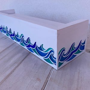 Peut inclure: Une étagère rectangulaire en bois blanc ornée d'un motif peint représentant des vagues stylisées dans des tons bleu foncé, bleu clair et sarcelle sur les panneaux avant et latéraux. L'étagère possède une surface supérieure plate.