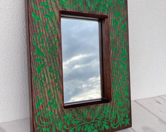 Op de afbeelding: Een houten spiegel met een groen bloemmotief dat in de lijst is gegraveerd.