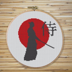 May include: Cross-stitch embroidery of a samurai silhouette standing in front of a red sun with a tree and Japanese characters in the background.