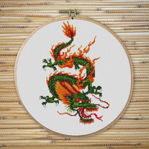Può includere: Ricamo a punto croce di un drago verde e arancione su tessuto bianco, incorniciato in un cerchio di legno. Il drago è raffigurato con dettagli intricati, tra cui una lingua rossa e accenti fiammeggianti. Lo sfondo è una trama naturale tessuta.