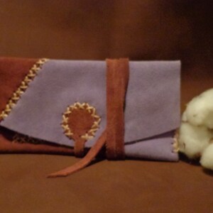 May include: A purple and brown suede leather pouch with a brown leather strap and a decorative stitching detail. The pouch is closed and sitting on a brown surface.