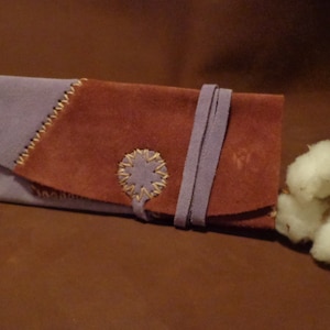 May include: A brown and purple leather pouch with a decorative stitch detail and a strap closure. The pouch is sitting on a brown surface.