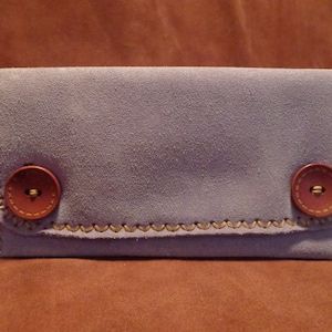 May include: A small, light purple suede pouch with two brown buttons. The pouch has a stitched edge.