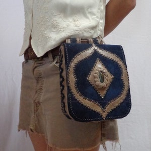 May include: A brown suede and blue leather fanny pack with a decorative diamond-shaped flap and a small, round, green gemstone in the center. The fanny pack is worn on a person's waist.