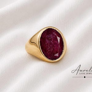 Könnte beinhalten: Ein goldener Siegelring mit einem großen, oval geschliffenen, tiefroten Rubin in der Mitte. Der Ring hat eine polierte, glatte Goldschiene. Der Text "Aurelia LUXE JEWELS" ist in der unteren rechten Ecke des Bildes aufgedruckt.