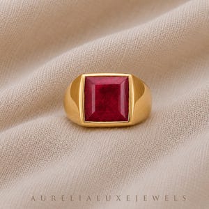 May include: A gold ring with a large, square, deep red gemstone. The ring has a thick band and a simple, elegant design. The ring is set against a neutral background. The text "AURELIA LUXE JEWELS" is at the bottom of the image.