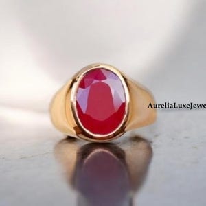 Puede incluir: Un anillo de oro con una gran piedra roja de forma ovalada. El anillo está engastado en un estilo simple y clásico.