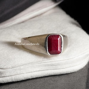 Puede incluir: Un anillo de plata con una piedra preciosa roja de forma rectangular. El anillo está sobre una superficie blanca.