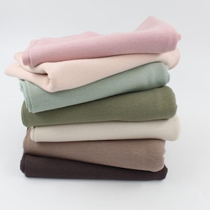 Könnte beinhalten: Ein Stapel von sieben Stoffproben in verschiedenen Farben, darunter Rosa, Beige, Grün, Oliv, Creme, Braun und Dunkelbraun. Der Stoff ist weich und gestrickt.