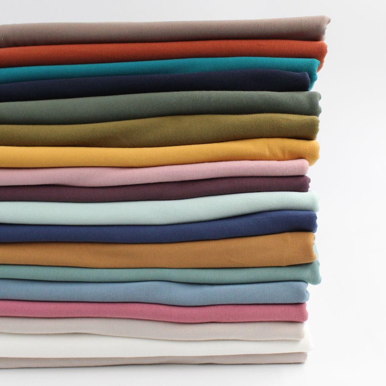 May include: A stack of folded fabric in various colors, including brown, teal, navy, olive green, mustard yellow, and pink. The fabric appears to be a soft material, possibly cotton or linen, suitable for sewing or crafting projects.