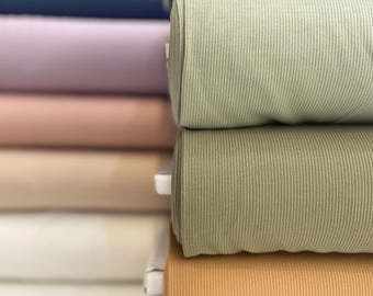 May include: A vertical stack of folded fabric bolts featuring a ribbed texture. The colors include teal, sage green, olive green, mustard yellow, and muted brown. Each bolt is neatly rolled and layered, showcasing the consistent vertical ribbing pattern.