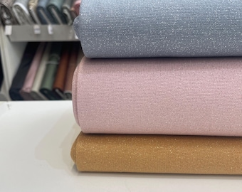 Puede incluir: Tres rollos de tela apilados con un sutil brillo metálico. El rollo superior es azul claro, el central es rosa suave y el inferior es de un color amarillo mostaza cálido. Cada rollo muestra una textura tejida fina y consistente en toda su superficie.