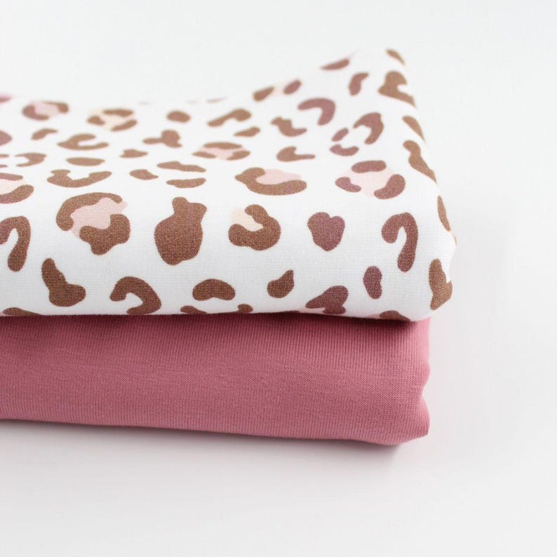Zwei aufeinandergelegte Jersey-Stoffe. Der obere Stoff hat ein modernes Leopardenmuster in Braun- und Rosatönen auf weißem Hintergrund. Der untere Stoff ist ein einfarbiger, zart rosafarbener Jersey.
