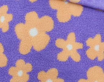 May include: A plush, textured fabric featuring a repeating pattern of light orange flowers with white centers set against a vibrant purple background. The material has a soft, fuzzy surface with a folded edge showing the same floral design.