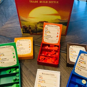 Könnte beinhalten: Ein Catan-Brettspiel-Set mit fünf verschiedenen farbigen Boxen, die Holzteile und Karten enthalten. Jede Box enthält eine Karte, die die Baukosten für Straßen, Siedlungen, Städte und Entwicklungskarten zeigt. Die Karten sind mit den benötigten Ressourcentypen für den Bau jedes Elements beschriftet.