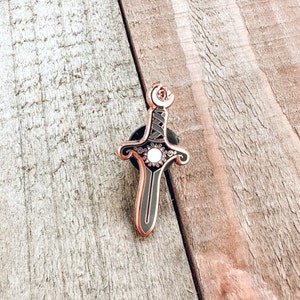 May include: Rose gold enamel pin of a sword with a black and white design. The sword has a crescent moon at the top and a sun with a white center in the middle.