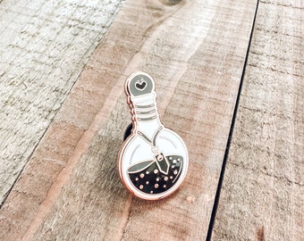 May include: A rose gold enamel pin of a potion bottle with a black liquid and white glitter inside. The bottle has a white stopper with a small heart on top and a white string with a charm hanging from the neck.