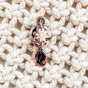 May include: Rose gold enamel pin featuring a black cat paw holding a white flower with a yellow center.