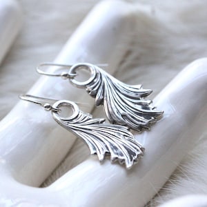 A pair of sterling silver dangle earrings with detailed leaf-like designs, resting on a white ceramic hand display. The earrings have intricate curves and texture, resembling flowing leaves or feathers.