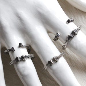 May include: A collection of silver-toned rings with an organic, textured design. The rings are displayed on a white hand-shaped form, showcasing their unique, branch-like appearance. The rings vary in size and style.