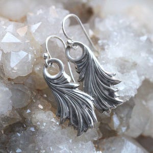 May include: A pair of silver earrings with a delicate, feather-like design. The earrings are displayed on a bed of white crystals.