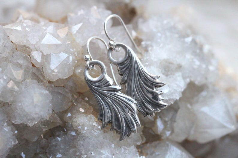 May include: A pair of silver earrings with a delicate, feather-like design. The earrings are displayed on a bed of white crystals.