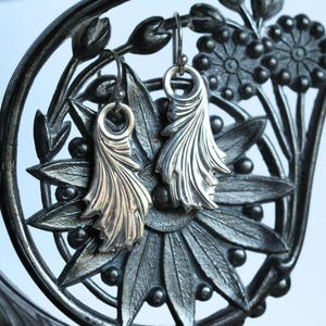 May include: A pair of silver earrings with a delicate, leafy design. The earrings are hanging from a black metal stand with floral details.
