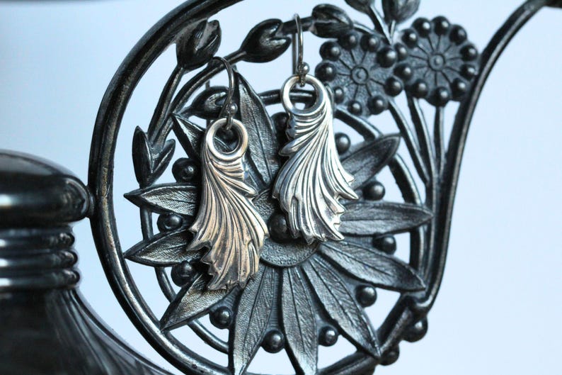 May include: A pair of silver earrings with a delicate, leafy design. The earrings are hanging from a black metal stand with floral details.