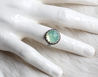 May include: A silver ring with a round, light green gemstone set in a silver bezel. The bezel has a decorative, scalloped edge.