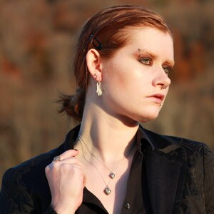 May include: A woman with red hair wearing a black blazer with a silver necklace and earrings. She has a nose ring and a silver stud earring in her left ear.
