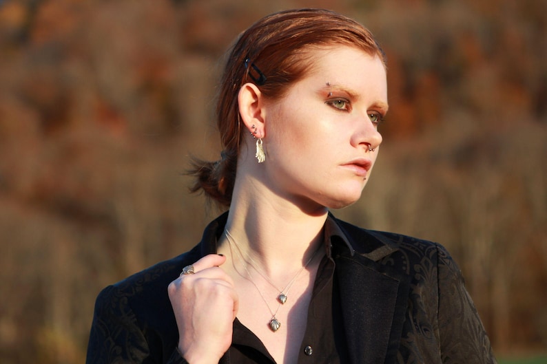 May include: A woman with red hair wearing a black blazer with a silver necklace and earrings. She has a nose ring and a silver stud earring in her left ear.