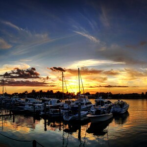 May include: A marina at sunset with a variety of boats docked. The sky is a mix of blue, orange, and yellow with clouds. The water is calm and reflecting the colors of the sky.