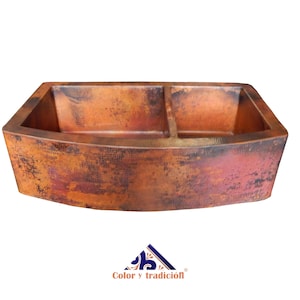 May include: A double-bowl copper kitchen sink with a farmhouse style design. The sink has a hammered finish and a warm, rustic color.