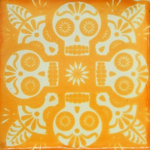 May include: A yellow ceramic tile with a white design of four stylized skulls with floral accents. The skulls have large eyes and a simple nose and mouth.
