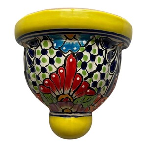 May include: A hand-painted ceramic wall planter with a vibrant floral design. The planter features a yellow rim and base, with a colorful pattern of red, blue, green, and white flowers and leaves. The planter is approximately 8 inches wide and 7 inches tall.
