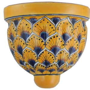 May include: A yellow ceramic wall planter with a decorative pattern of stylized blue and yellow fan-like shapes. The planter has a rounded bottom and a small, rounded base. The rim is a darker yellow with a thin blue stripe.