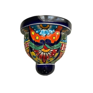 May include: A hand-painted ceramic wall pocket with a vibrant, colorful design. The piece features a deep blue rim and base, with a central design of orange, yellow, green, and red floral patterns. The wall pocket is designed to hold small plants or decorative items.