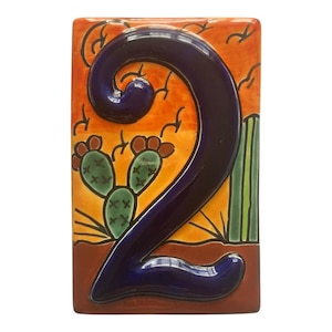May include: Rectangular ceramic tile featuring a large, glossy, dark blue number 2. The background is a desert scene with cacti, a setting sun, and a brown base. The tile has a glossy finish and is likely for a house number.