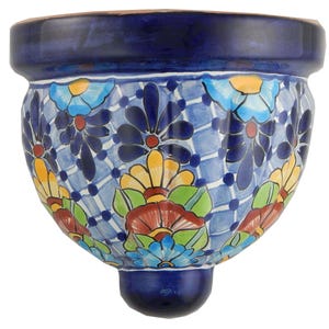 May include: A hand-painted, ceramic wall planter with a vibrant floral design. The planter features a deep blue rim and base, with a light blue background adorned with colorful flowers in shades of yellow, red, and blue. The planter has a rounded shape and a small, rounded base.