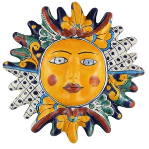 May include: A ceramic sun wall plaque featuring a central yellow face with blue eyes and red cheeks. The sun rays are decorated with intricate patterns in blue, green, yellow, and white, including floral motifs and a blue-dotted grid design.