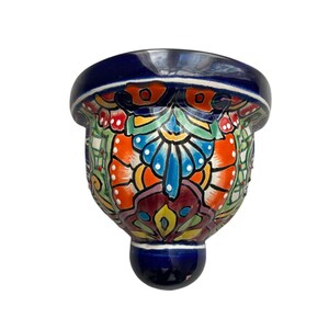 May include: A hand-painted ceramic wall pocket with a vibrant floral design. The piece features a deep blue rim and base, with a colorful pattern of orange, blue, green, and red. This decorative item is suitable for displaying small plants or other decorative items.